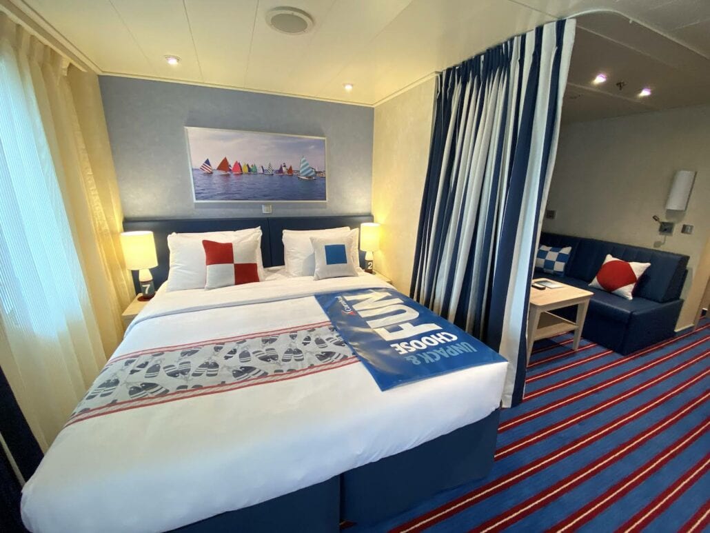 Step inside a Carnival Panorama cruise ship cabin where a neatly made double bed with nautical-themed bedding awaits. A blue sofa, small coffee table, and sailboat artwork enhance the charm. Striped carpet and curtains complement the soft lighting from bedside lamps in this cozy retreat.