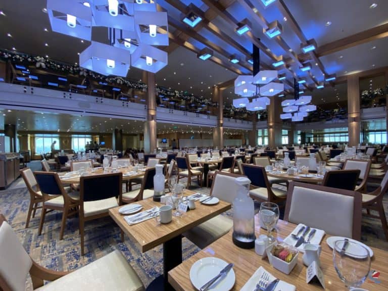 A spacious, brightly-lit restaurant with neatly arranged tables set for dining. Featuring a special New Year's Eve Menu, tables boast white dinnerware, glasses, and cutlery. Modern light fixtures adorn the ceiling, while decorative plants grace the upper level balcony.