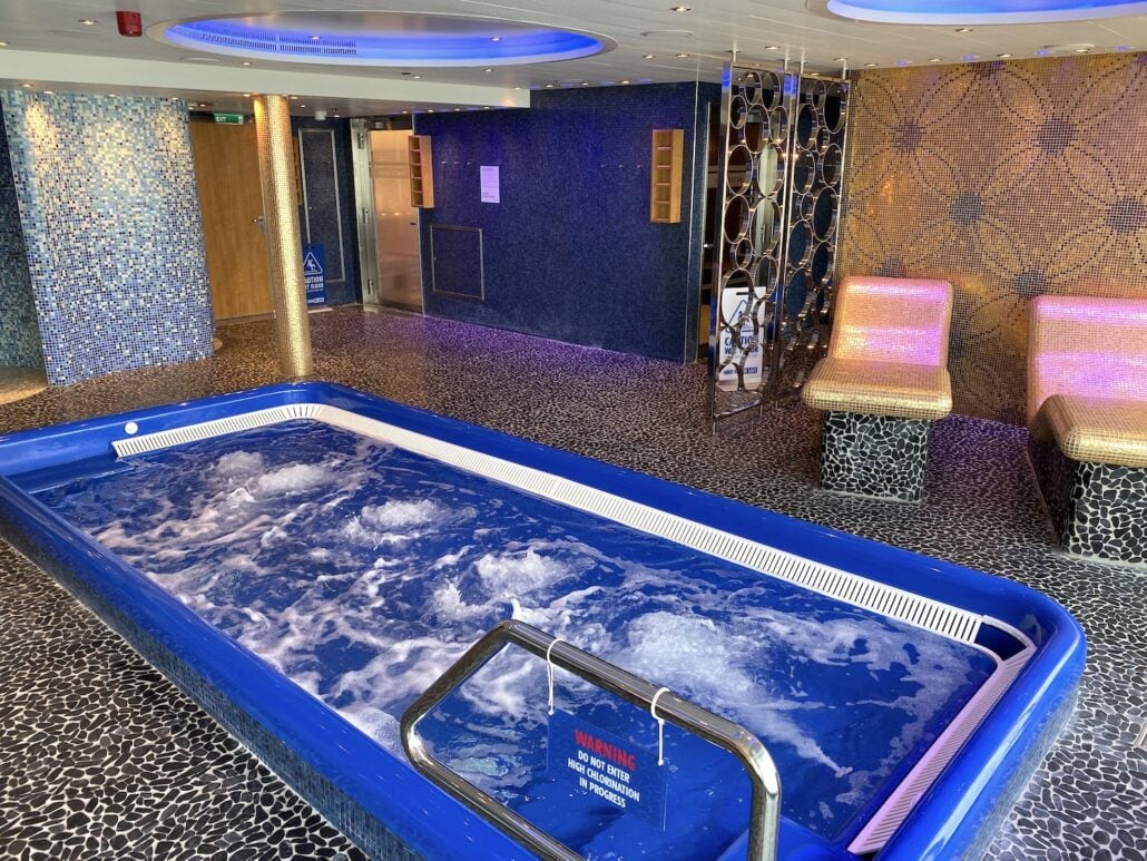 The indoor spa area aboard the Carnival Panorama boasts a rectangular blue jacuzzi with bubbling water and mosaic tile walls. Two cushioned seats nearby invite relaxation, while a metal handrail lines the edge. A "Warning" sign playfully floats in the water, adding a distinctive touch to this serene setting.