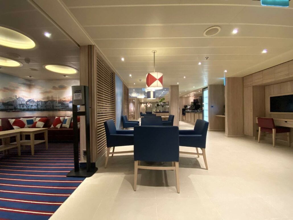 The modern lounge area on the Carnival Panorama boasts blue cushioned chairs around a wooden table, perfect for relaxing after a day of ship review. With wooden accents, patterned carpet, and a wall mural, the space is softly lit by a red and white pendant light.