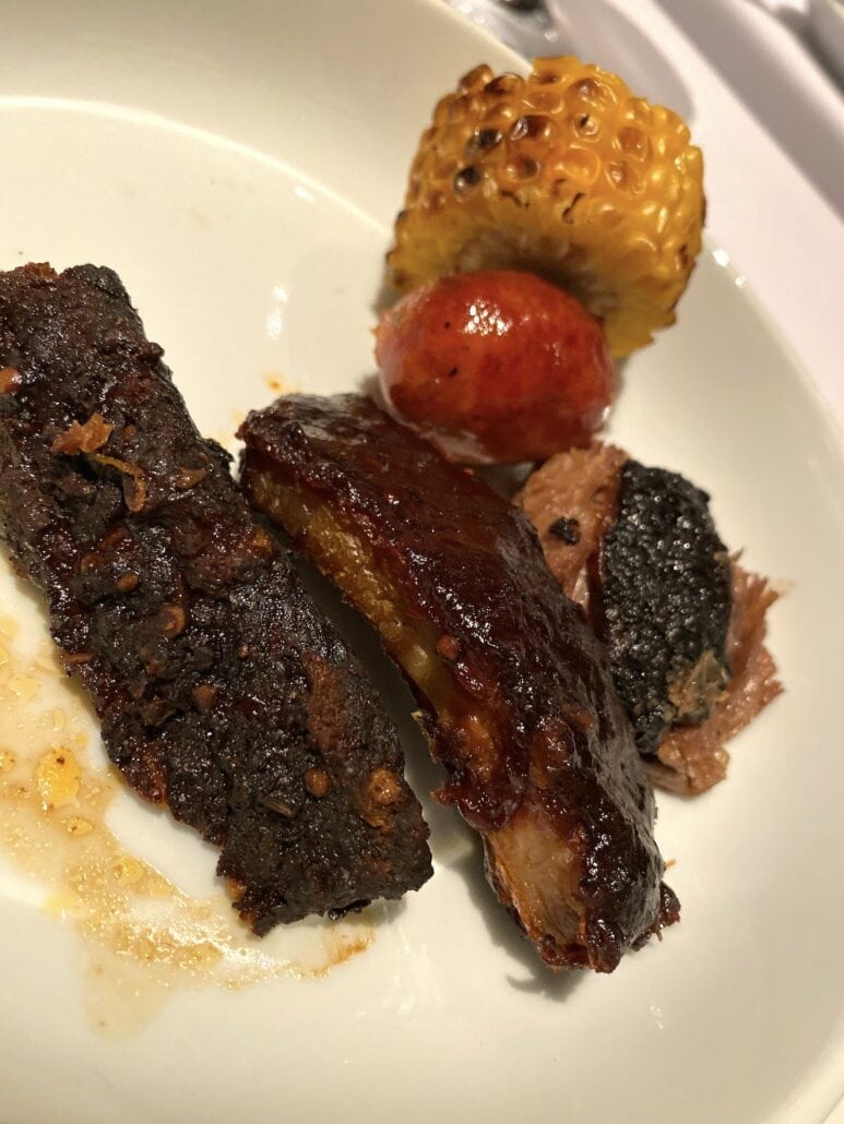 Plate with charred corn on the cob, a small roasted tomato, and glazed rib pieces captures the flavors you might discover on a culinary exploration aboard Carnival Panorama. The ribs boast a caramelized exterior, perfectly paired with the vibrant corn and juicy tomato for a delightful dining experience.
