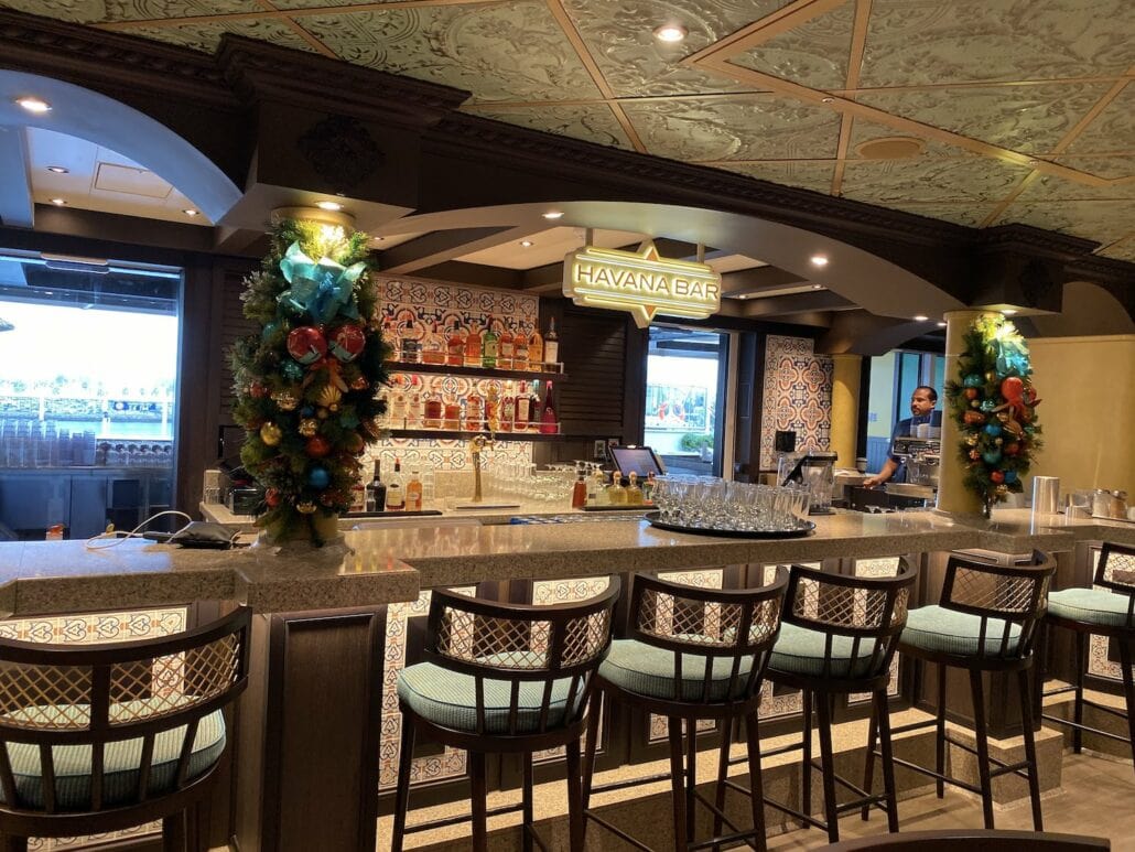 A spacious bar with a counter adorned with tropical-themed tiles is shown, reminiscent of the festive vibe aboard Carnival Panorama. Several bar stools surround the counter, while decorative wreaths hang near the entrance. A sign reads "Havana Bar," capturing a scene perfect for travel photos.
