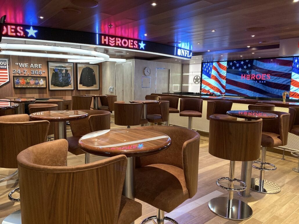 A modern bar on the Carnival Panorama, with wooden tables and brown stools. The walls feature American flags and "HEROES SPORTS BAR" signage, while display cases showcase military uniforms. LED lights adorn the ceiling, and flat-screen TVs provide entertainment—perfect for capturing memorable photos.