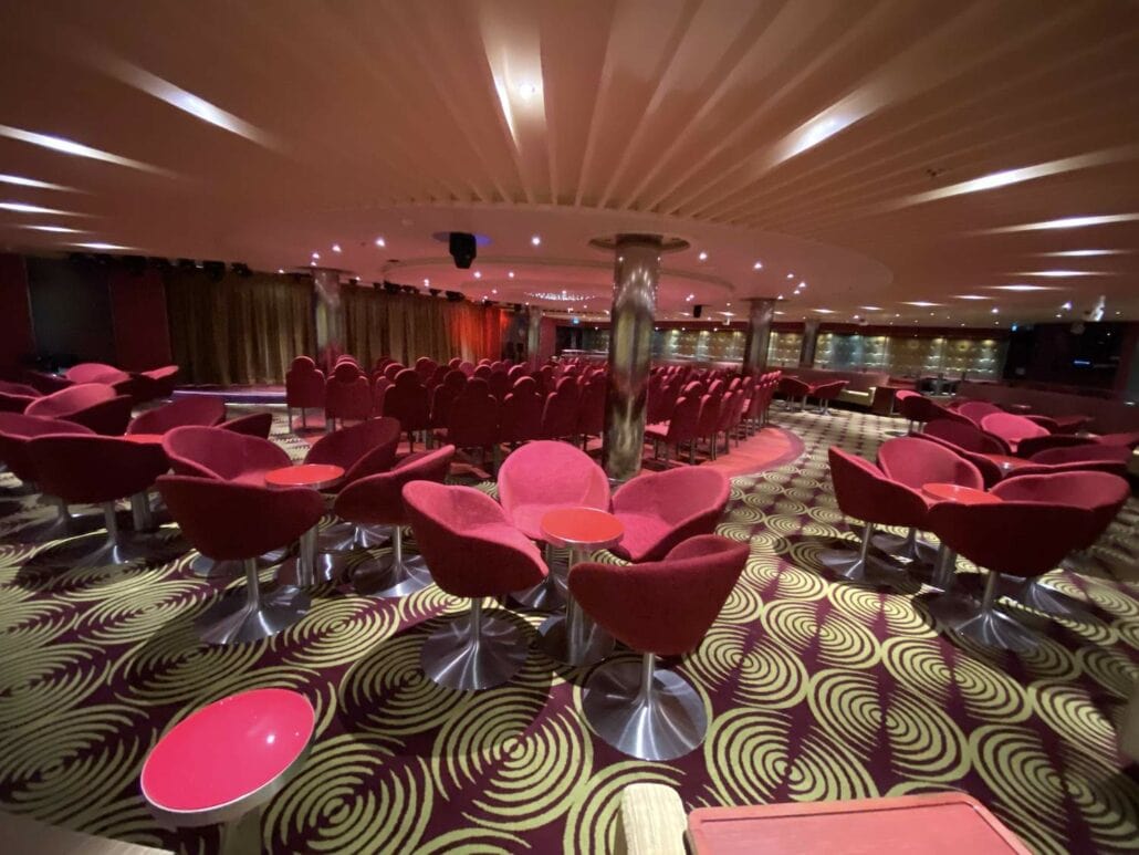 The dimly lit lounge aboard the Carnival Panorama features rows of red cushioned chairs and small round tables. A swirling green and purple carpet extends across the space, leading to a stage area under a low ceiling with spotlights—perfect for capturing unforgettable ship photos.