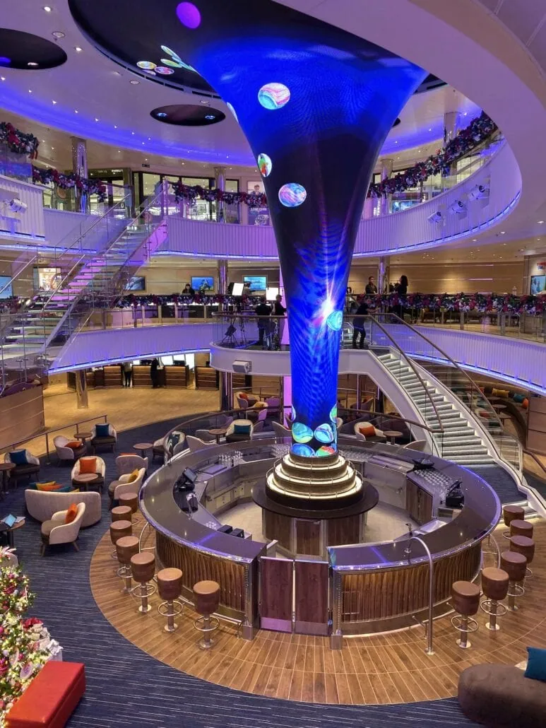 Step into the modern multi-level atrium of Carnival Panorama, where a large, illuminated sculpture commands attention. Curving staircases lead to seating areas with colorful chairs and a circular bar. Soft purple lighting enhances the elegant ambiance, making it perfect for capturing stunning photos.