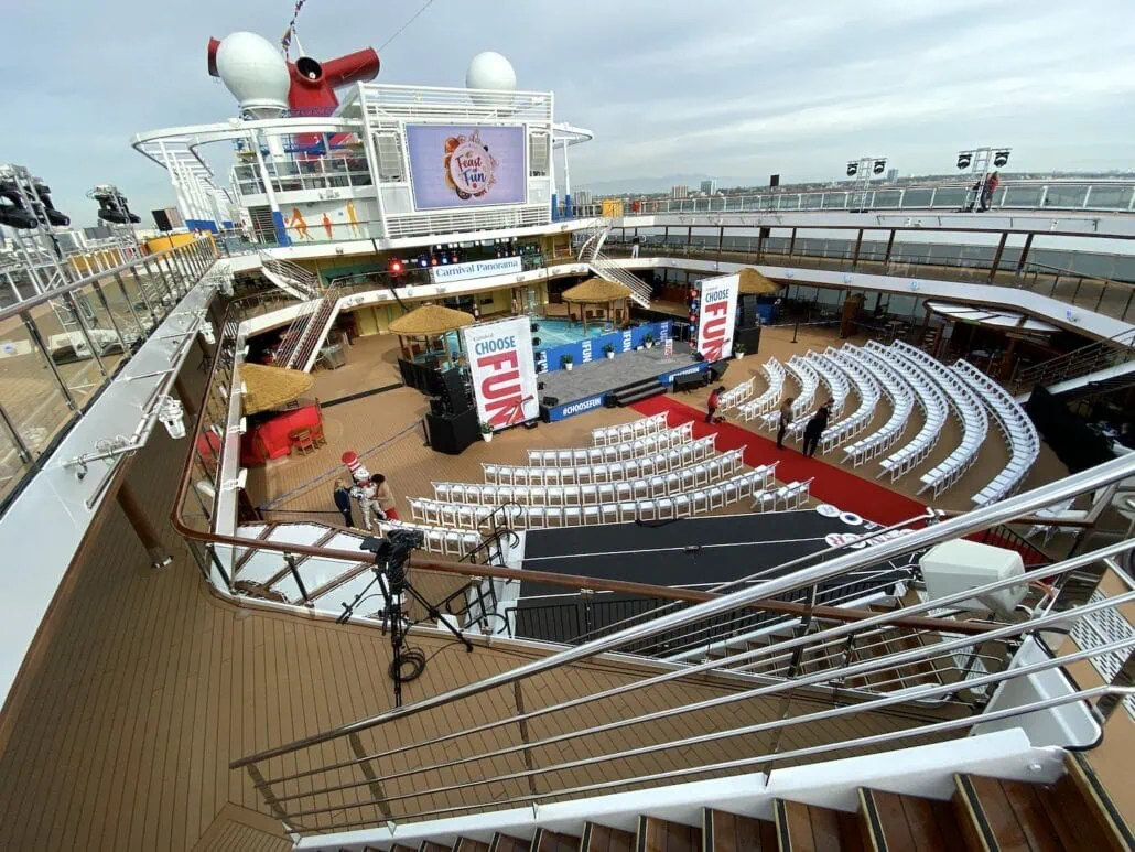 A cruise ship deck is arranged for an outdoor event, perfect for a Carnival Panorama experience. Rows of white chairs face a stage with a "Carnival Fun" screen. A red carpet lines the aisles, while the ship's structure and cityscape provide a picturesque backdrop worth capturing in photos.