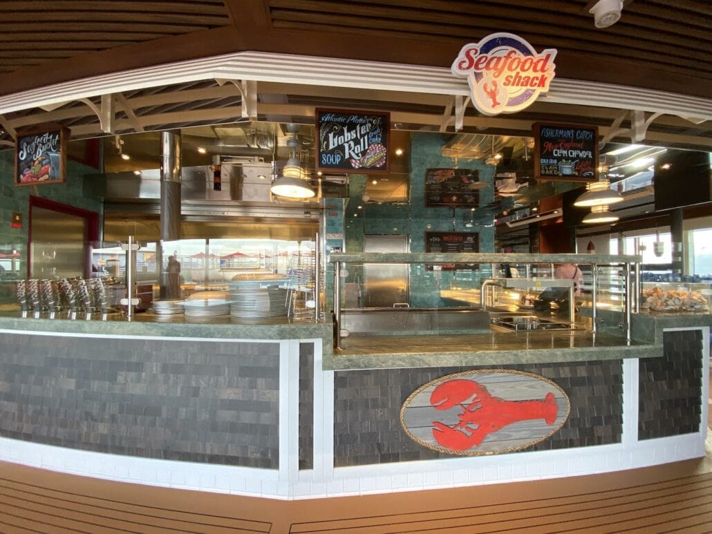 A seafood restaurant counter on the Carnival Panorama features a lobster logo and a "Seafood Shack" sign. Displaying photos of tempting items like lobster rolls and fries, the menu sits above busy kitchen equipment where a chef expertly prepares food for eager guests.