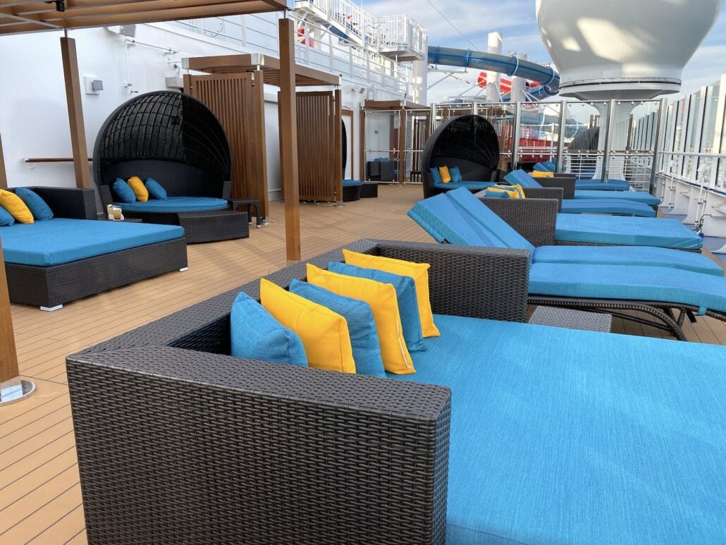 Aboard the Carnival Panorama, the cruise ship deck features wicker lounge chairs with blue cushions and yellow pillows for a pop of color. Egg-shaped chairs and red slides add a playful vibe against wooden flooring, all captured in stunning photos beneath a partially cloudy sky.
