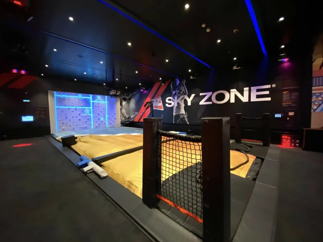 The "Sky Zone" indoor trampoline park onboard Carnival Panorama features padded flooring, a wrestling ring, and a climbing wall illuminated in blue. Perfect for both kids and adults, it's ideal for photos. The dim lighting is enhanced by spotlights, creating a playful atmosphere on the ship.