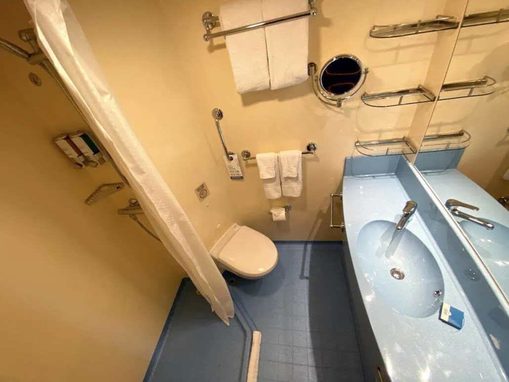 A small bathroom aboard the Carnival Panorama features a toilet, sink, and several towel racks. A round porthole window above the toilet and a blue floor adds to its nautical charm. The sink area boasts a large mirror and soap dispenser, perfect for a refreshing sea voyage experience.