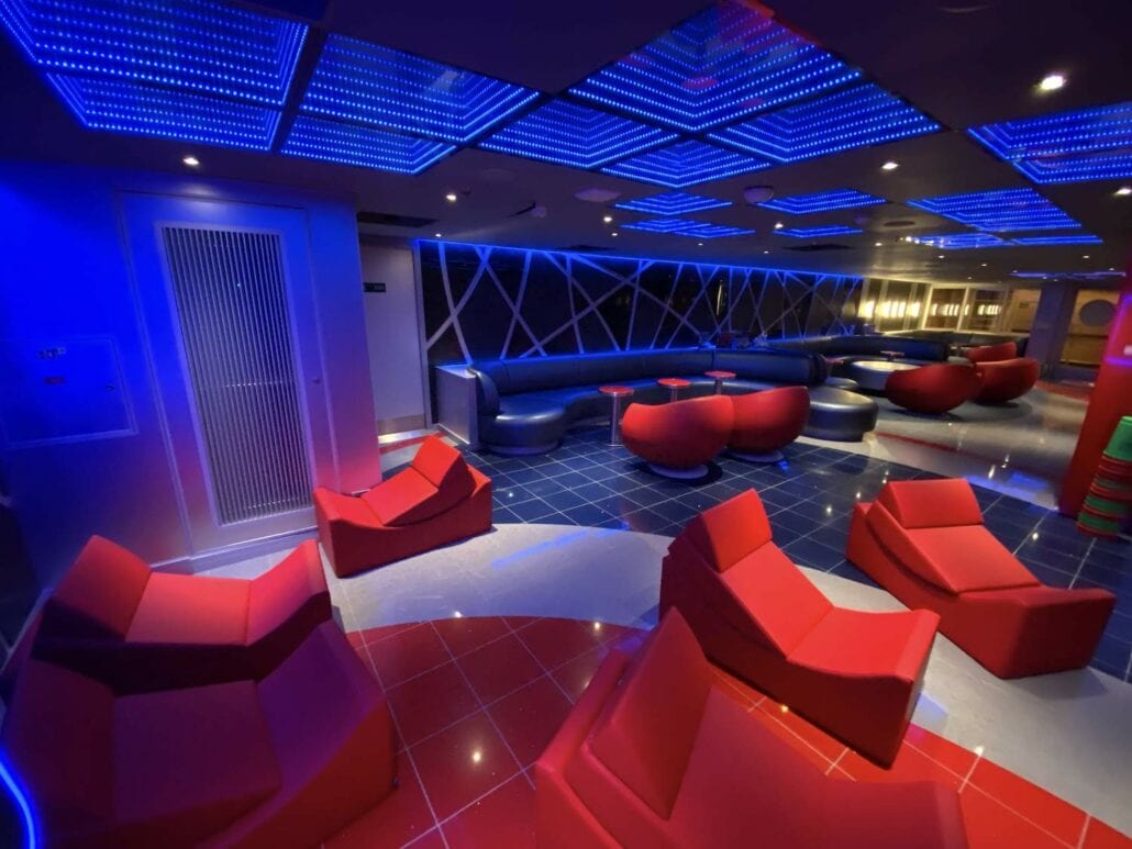 The image, likely from a Carnival Panorama ship review, showcases a modern lounge area with red and blue seating. The floor is tiled in those hues, while the ceiling boasts blue LED lighting. Futuristic chairs and round tables complement the long cushioned bench in the background, perfectly captured in these photos.