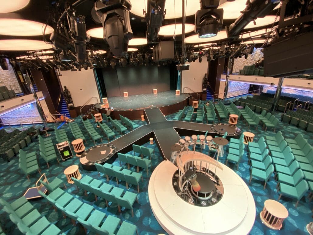 A theater aboard the Carnival Panorama features a circular stage with intimate seating arranged around it, creating the perfect ambiance for capturing unforgettable photos. Adorned with teal upholstery and decorative wall patterns, it's an inviting space where performances come to life under the glow of elegant lighting fixtures.
