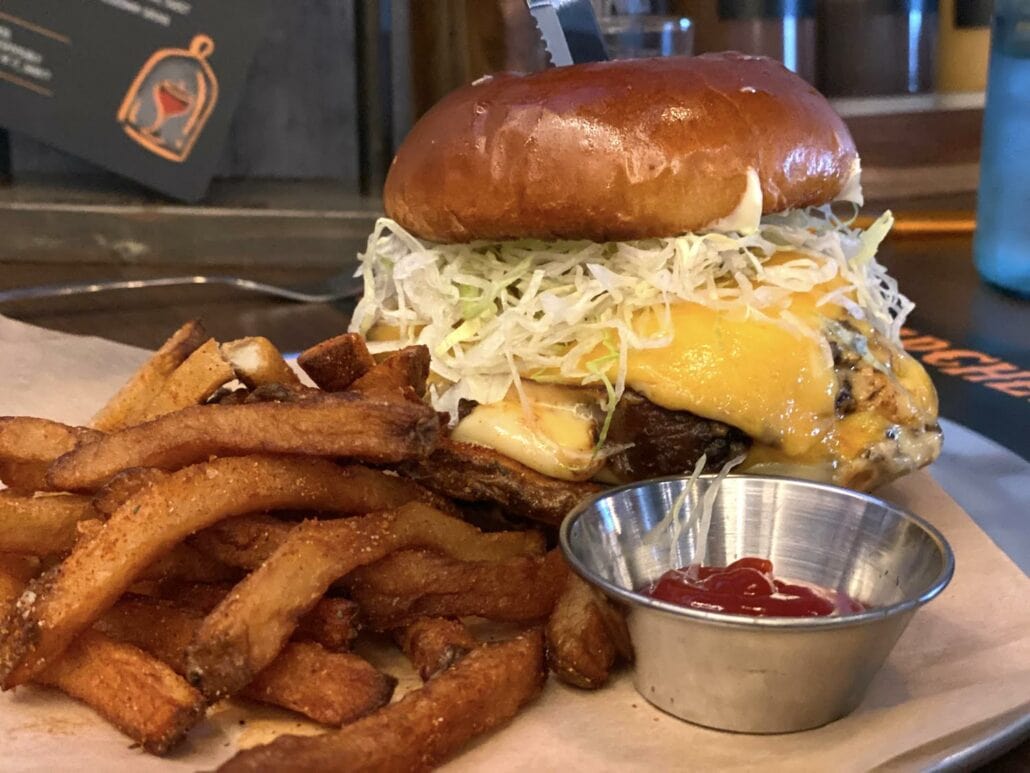 A cheeseburger topped with shredded lettuce and a knife in the bun, accompanied by a side of seasoned fries awaits on a paper-lined tray. This picture-perfect meal, resembling delights found on Carnival Panorama, includes a small metal cup of ketchup beside the fries.