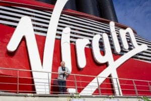 A person stands on a balcony in front of a red structure with the large white word "Virgin" prominently displayed, celebrating the completion of Virgin Voyages' first cruise ship. Black vertical elements are visible at the top against a blue sky background. The person is wearing a light jacket and pants.
