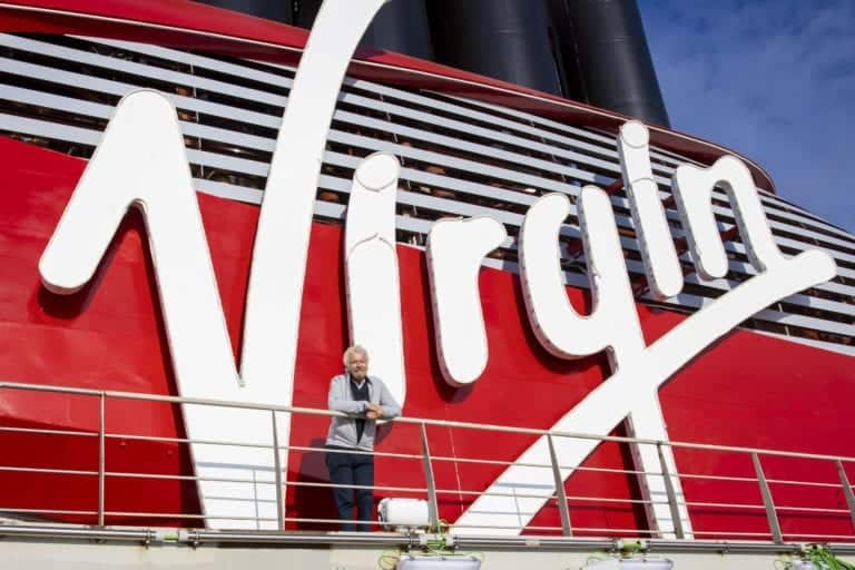 A person stands on a balcony in front of a red structure with the large white word "Virgin" prominently displayed, celebrating the completion of Virgin Voyages' first cruise ship. Black vertical elements are visible at the top against a blue sky background. The person is wearing a light jacket and pants.