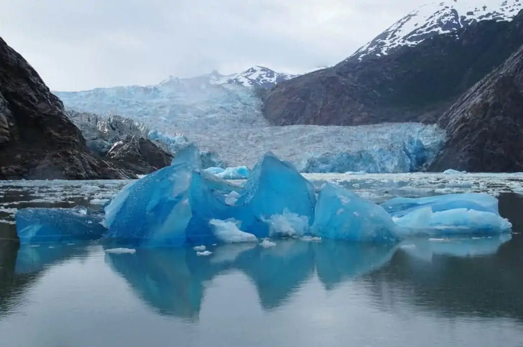 A bright blue iceberg floats in calm water, with Alaska's mountain range and snowy peaks in the background. The sky is overcast, and the Sawyer Glaciers in the distance blend seamlessly into the mountains, creating a breathtaking scene perfect for a memorable cruise.