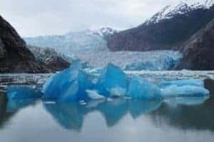 A vibrant blue iceberg floats in calm water with the majestic Sawyer Glaciers and snow-capped mountains in the background. The Alaskan sky is overcast, casting a cool tone over the serene landscape, making it a captivating sight on any cruise.