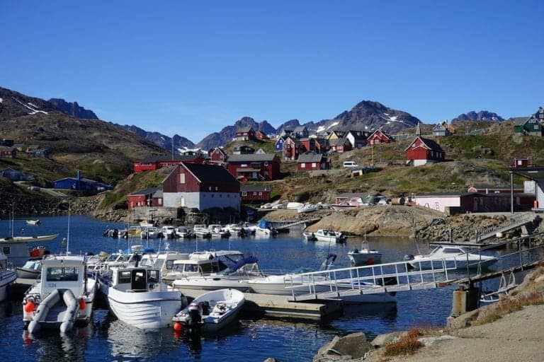 A small harbor with several white boats docked at a pier on a clear day. Red and white buildings and houses dot the hill overlooking the harbor. The background features rugged, rocky mountains against a bright blue sky, perfect for Destination Cruises seeking picturesque views.
