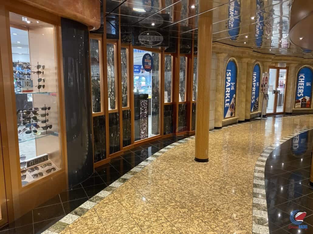 A corridor with a glossy black and tan floor in a shopping area features various storefronts. One store has sunglasses on display in a glass case, while another showcases a vibrant Carnival Pride tour photo gallery. The ceiling is reflective, adorned with wooden columns, and blue signs with text.