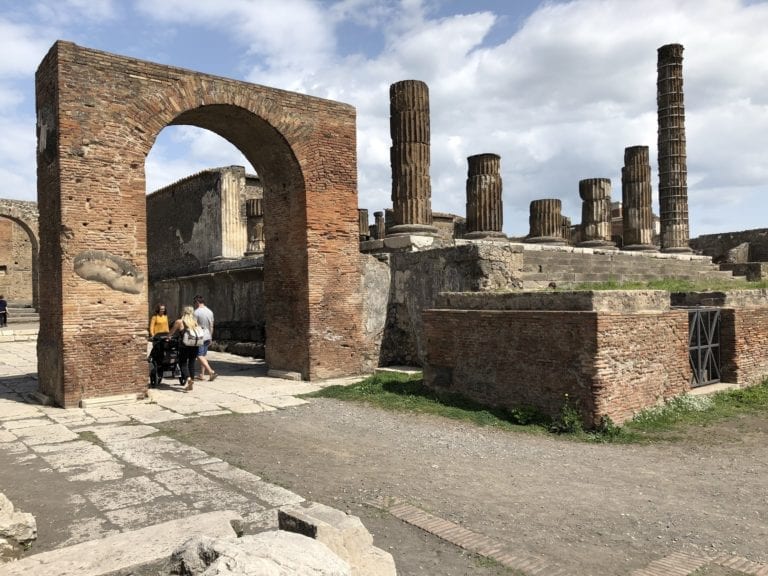An ancient ruin site in Naples with several stone columns and a large, arched entrance made of bricks. There are a few visitors in casual clothing, including a person pushing a stroller and two others walking nearby. The sky is partly cloudy, adding charm to this popular stop on Italy shore excursions.