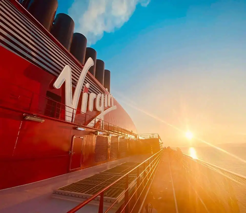 A photo of the side of a Virgin cruise ship at sunrise or sunset. The ship, painted in red with the word "Virgin" in white letters, has a stack of black smokestacks emitting light smoke. The sun is low on the horizon, illuminating the scene like a reward for every good deed done.