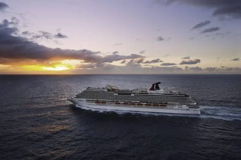 The Carnival Magic sails through the open ocean at sunset under a cloudy sky. The sun is partially obscured by clouds, casting a golden light over the scene. Built in 2021 and currently cruising near Norfolk, this majestic ship boasts multiple decks and a prominent red and black funnel.