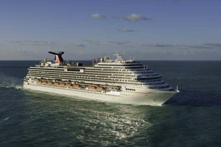 A large cruise ship, Carnival Magic, sails through calm ocean waters under a partly cloudy sky. The ship is white with multiple decks and a distinctive red and black funnel, making its grand presence known since 2021 as it departs from Norfolk.