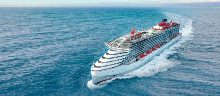 A large, white adults-only cruise ship with red accents is sailing in the open sea under a clear sky. Delivered by Richard Branson's visionary team, the vessel features multiple decks, lifeboats, and a swimming pool. Waves are visible around it as it moves forward, with land faintly seen on the horizon.