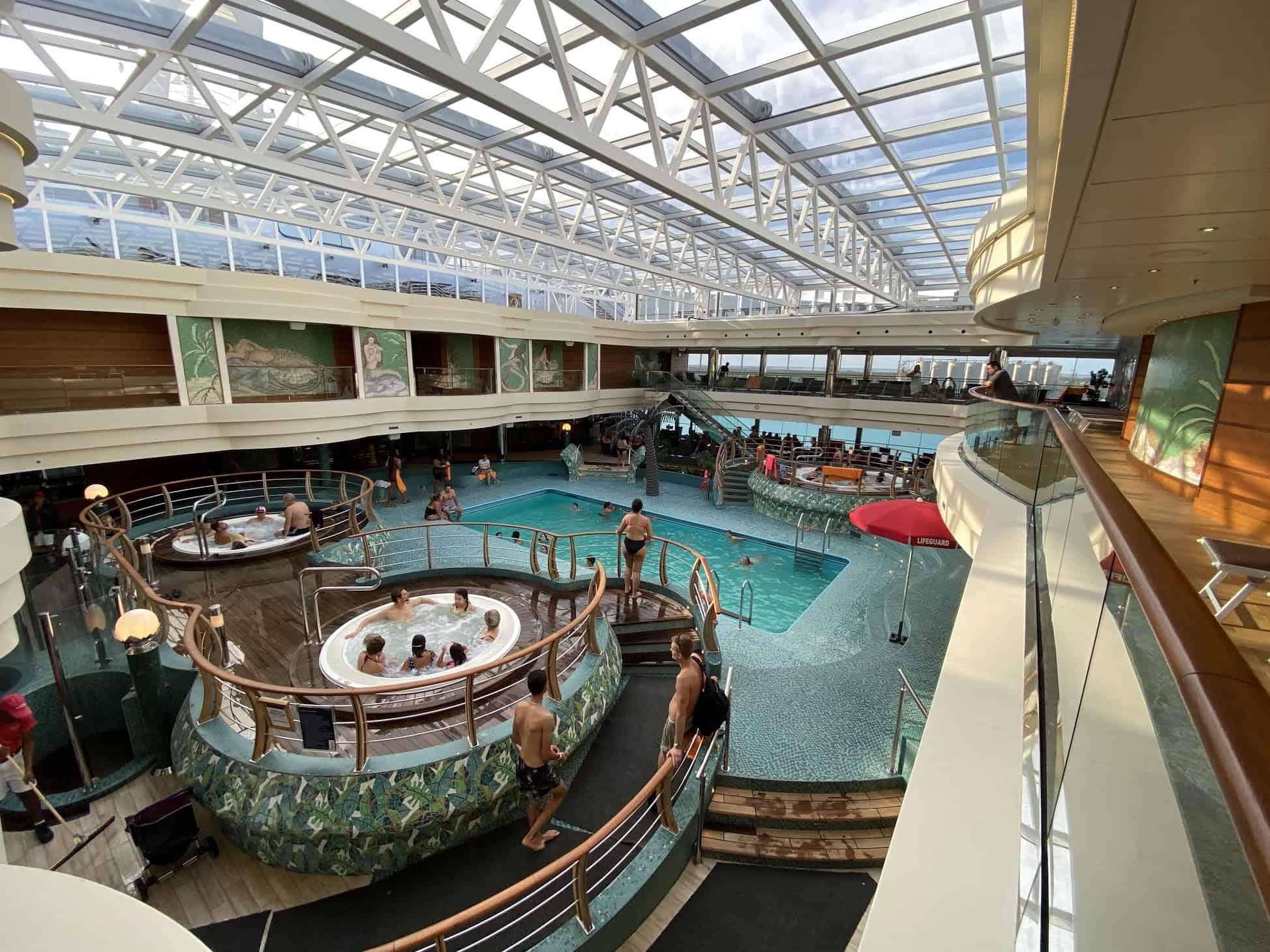 A large indoor swimming pool area with a transparent roof aboard the MSC Divina. Several people are in the main pool and hot tubs. A red umbrella and lounge chairs are visible. The area has multiple levels, with people walking and relaxing throughout the space, perfect for your photographic journey on MSC Cruises.