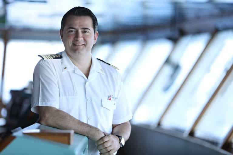 A senior officer dressed in a white naval uniform stands at the helm of the Carnival Radiance, with hands resting on a control panel. The background shows the windows of the ship's bridge and a blurry view of the sea, setting the stage for an upcoming Carnival adventure.