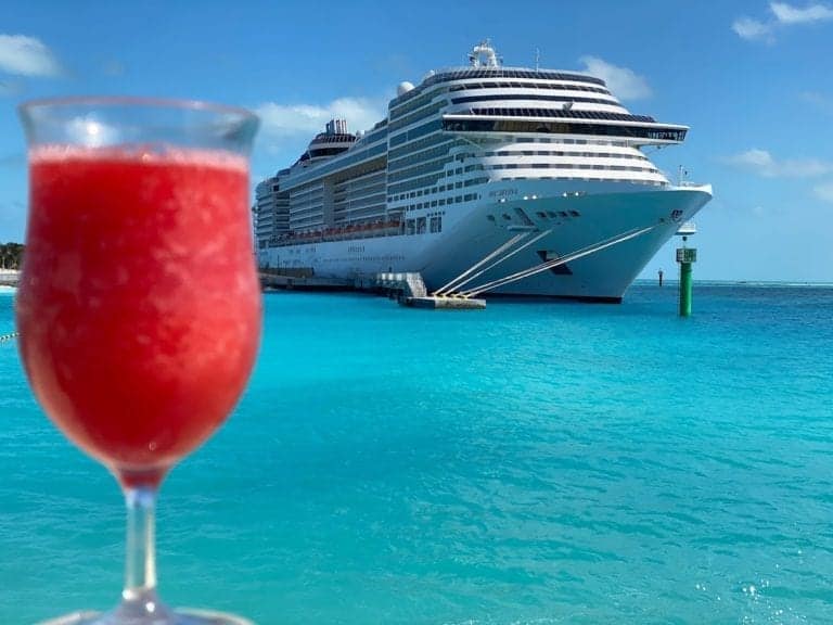 A large MSC cruise ship is anchored in clear turquoise water near the pristine Ocean Cay Marine Reserve under a blue sky. In the foreground, a red frozen drink in a tall glass is visible.