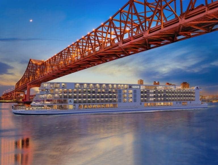 A large cruise ship reminiscent of Viking design sails on a calm Mississippi River at dusk, under a brightly lit steel bridge. The ship boasts multiple decks with numerous lit rooms and balconies. The sky is a gradient of blue and orange, with city buildings visible in the background.