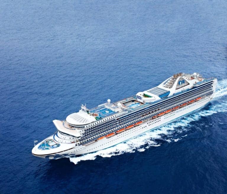 A large white cruise ship is sailing in the open ocean. The ship has multiple decks, some featuring pools and all have rows of blue-tinted windows. The sky is clear, and the ocean is calm with visible waves trailing the ship.