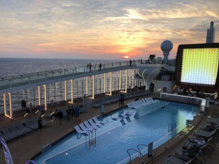A sunset view over Celebrity Edge's pool deck. The deck features lounge chairs, a large rectangular pool, a screen displaying a bright image, and several people walking around or sitting. The ocean is visible in the background under a cloudy sky—perfect for your next photo tour.