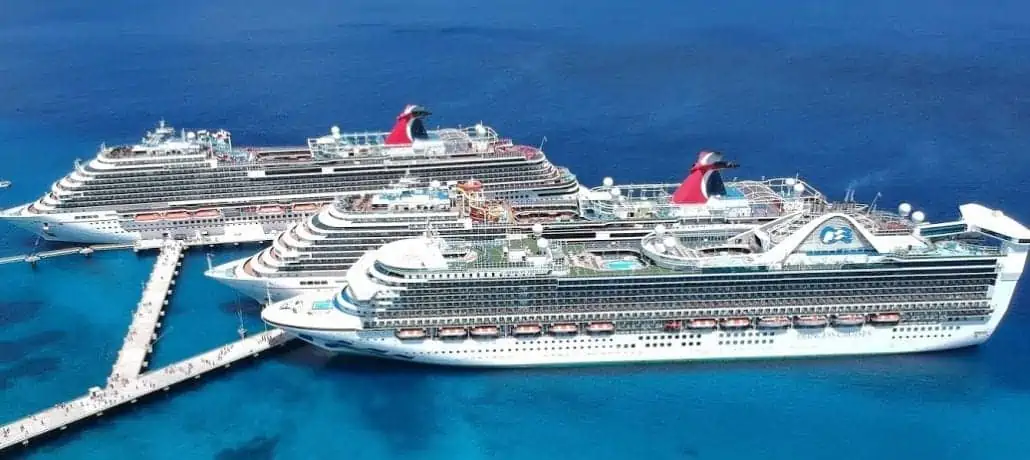 Three large cruise ships are docked side by side at a pier in clear, blue water. The white ships boast multiple decks filled with amenities, all adhering to high cleanliness standards. Viewed from above, passengers stroll along the pier against the backdrop of the expansive ocean—hallmarks of today's meticulous cruise industry.