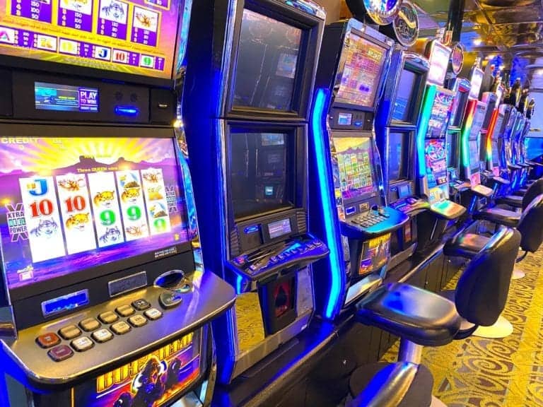 A row of brightly lit slot machines with various games onscreen is seen in a lively Florida casino ship environment. Empty black chairs are positioned in front of each machine, and the floor is carpeted with a patterned design, setting the stage for an exciting cruise gambling experience.