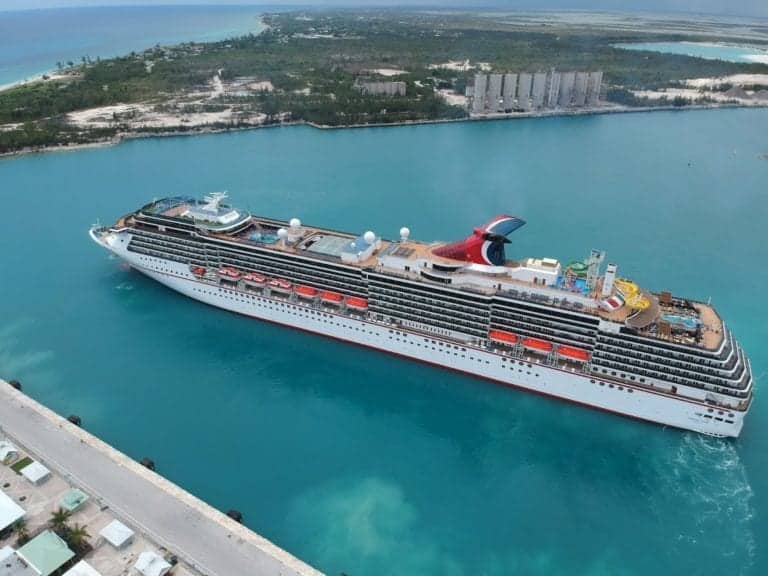An aerial view of a large cruise ship docked at Freeport with turquoise water. The vessel has multiple decks with lifeboats visible along the sides and various amenities on the top deck, including pools and seating areas. The coastline and buildings are in the background.