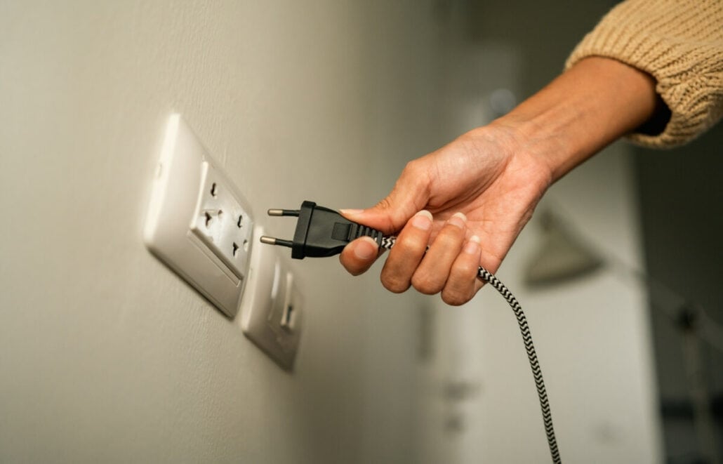 A person wearing a beige sweater carefully takes the steps to plug a black and white braided electrical cord into a wall socket, as if in preparation for the start of an exciting new cruise.