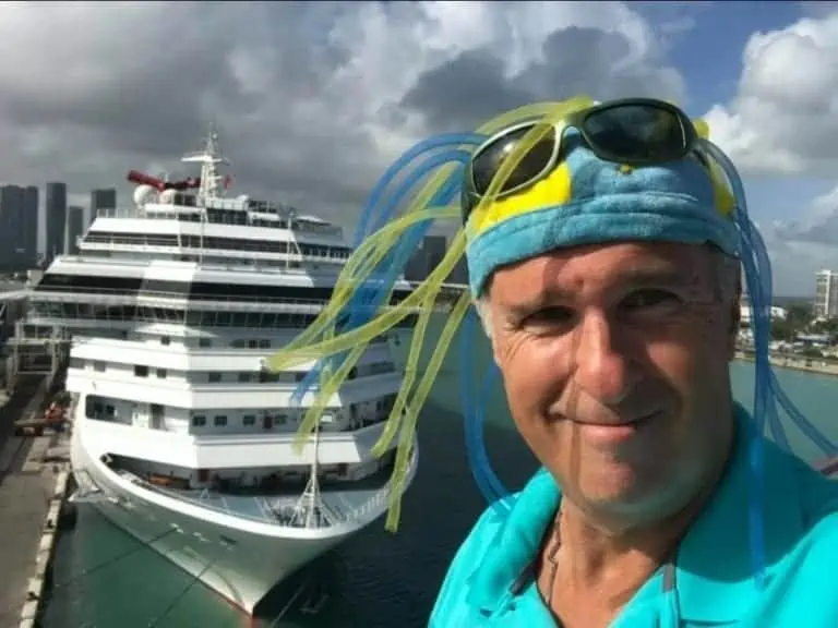A man, known as Stormin' Norm, wearing a blue and yellow hat with streamers and sunglasses stands in front of a large cruise ship docked at a port. The sky is cloudy, and city buildings are visible in the background. Smiling on the deck overlooking his favorite cruiser, he enjoys the moment in memory of past adventures.