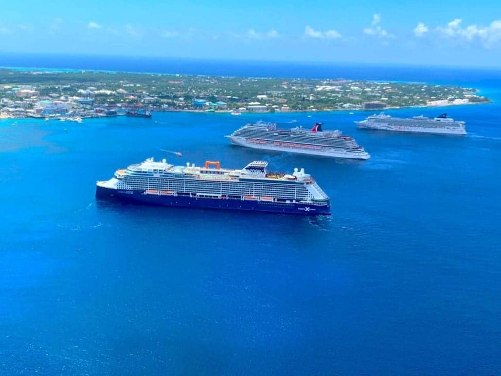 Aerial view of three large cruise ships anchored near a Caribbean island's coastline. The ships are in a calm blue sea, with a coastal town and green vegetation visible in the background. The sky is clear with a few scattered clouds, capturing the serene moment of 2021.