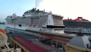 Two large cruise ships docked at a port, with the closer ship emitting white smoke from its funnel. The sky is clear, and some port facilities are visible in the foreground. One ship, associated with a prominent cruise line, has a white hull, while the other boasts black and red design as they return to service.