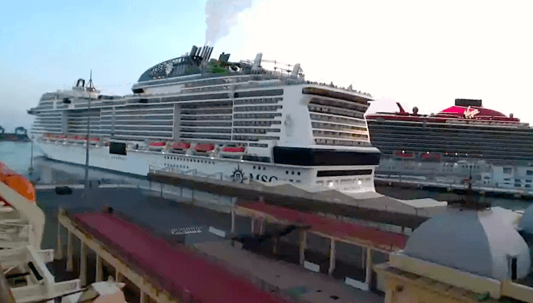Two large cruise ships docked at a port, with the closer ship emitting white smoke from its funnel. The sky is clear, and some port facilities are visible in the foreground. One ship, associated with a prominent cruise line, has a white hull, while the other boasts black and red design as they return to service.