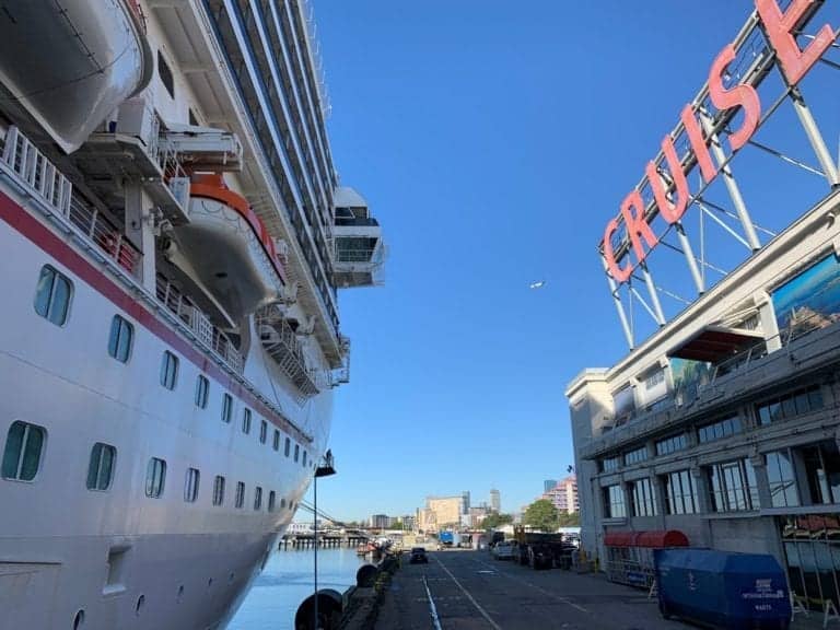 A large cruise ship docked peacefully at a port under the clear blue sky, reminiscent of serene European cities. To the right, a building adorned with a "CRUISE" sign stands proudly while a small aircraft soars above the sprawling cityscape in the background.