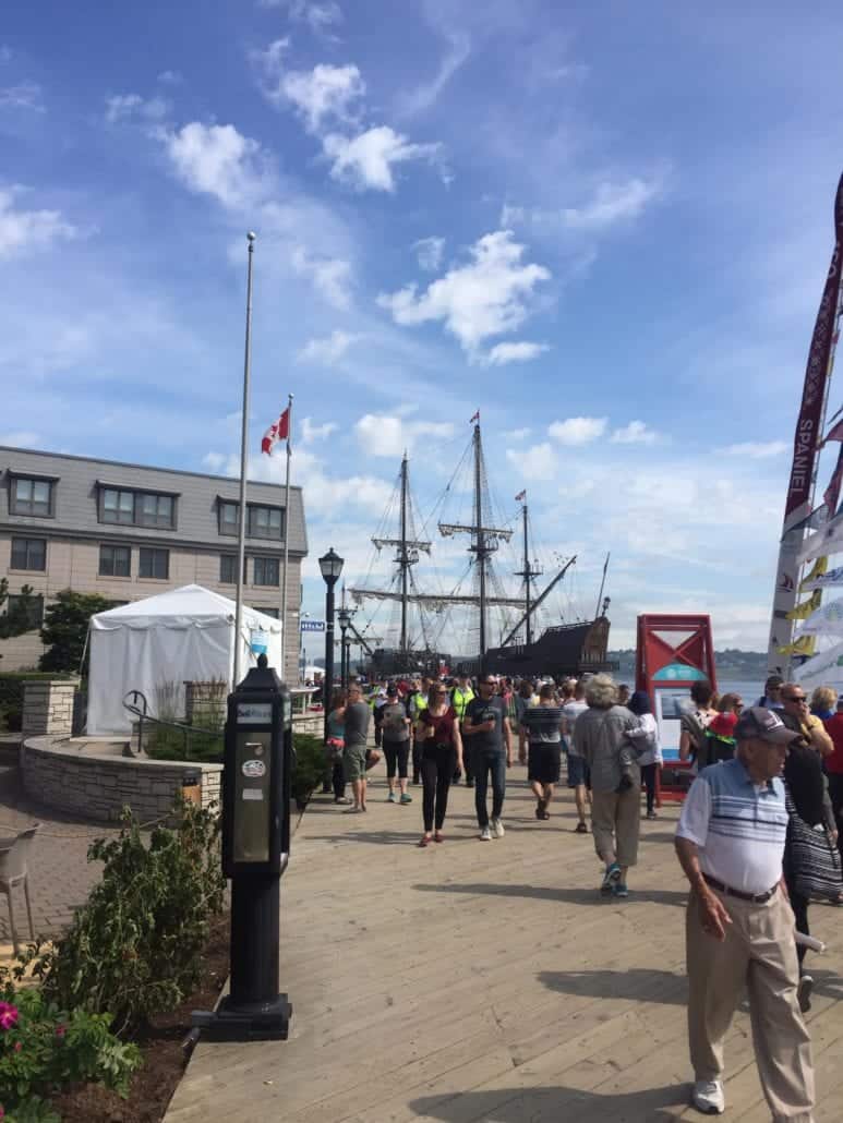 People walking on a waterfront boardwalk with an old-style sailing ship docked in the background. A Canadian flag flies on a pole to the left, and a modern building is visible in the background. The weather is sunny with a few clouds in the sky—perfect for exploring things to do at Halifax Cruise Port.