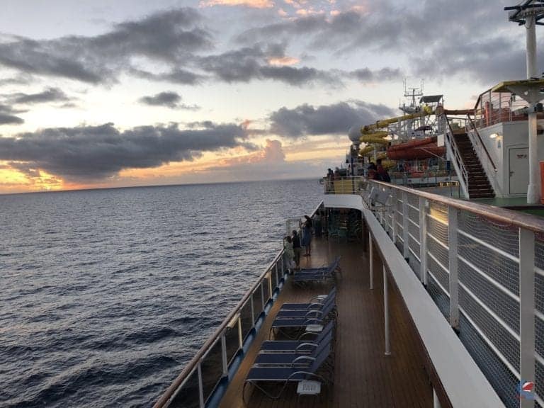 A cruise ship deck at sunset with lounge chairs arranged along the railing. The sky is partly cloudy with the sun setting on the horizon. Parts of a water slide are visible to the right, and a few people are seen on the deck near the railings.