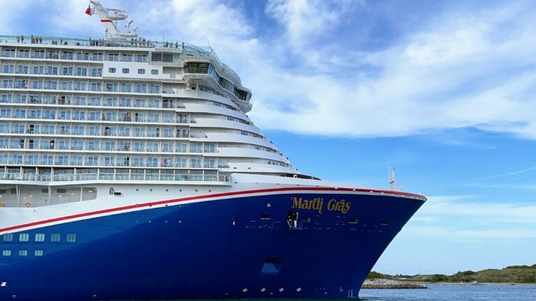 A large cruise ship named "Mardi Gras" is docked at Port Canaveral. The vessel boasts multiple decks with balconies and is painted predominantly blue with white and red accents. The sky is mostly clear with a few clouds, and some greenery is visible in the background.