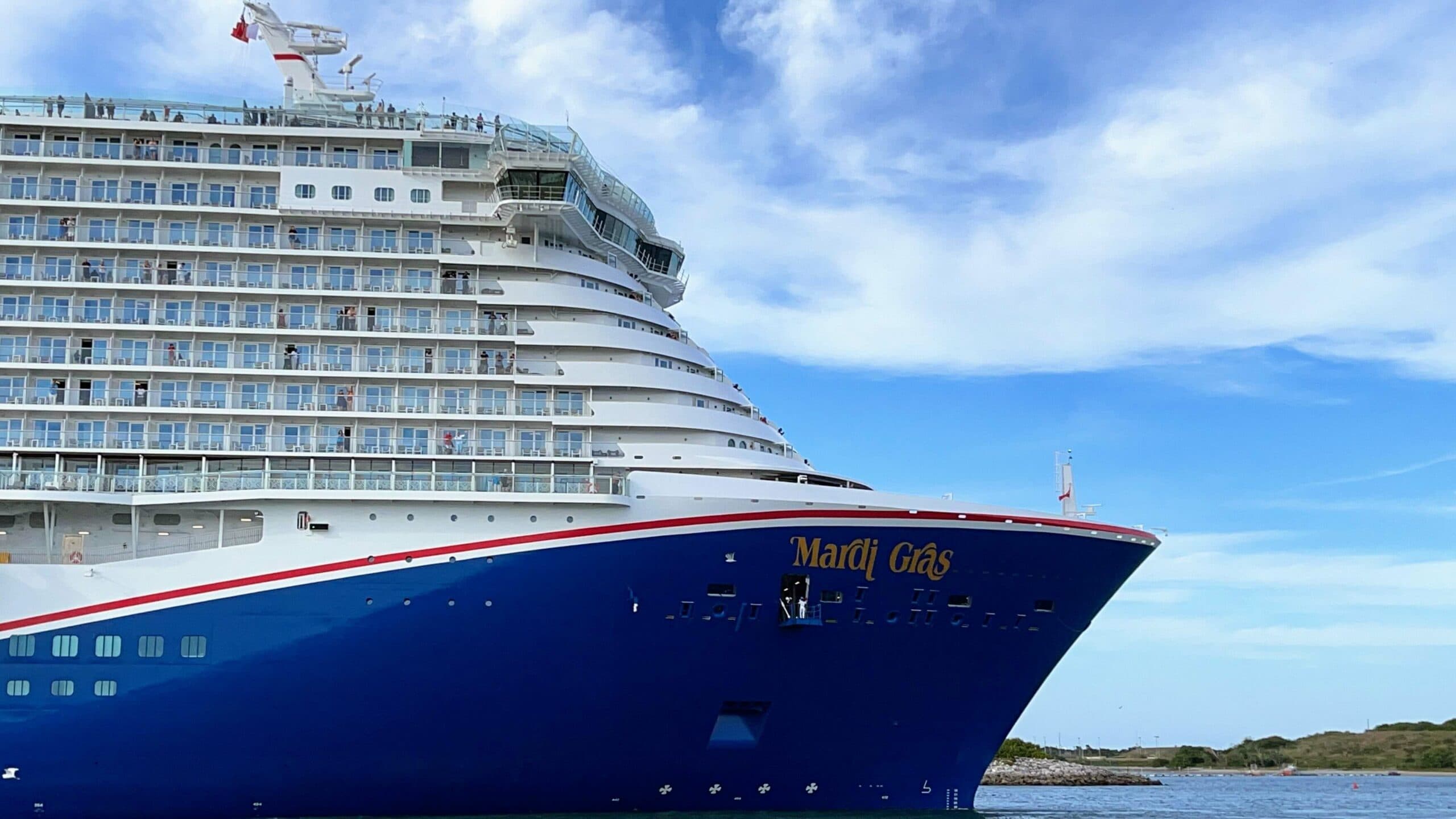 A large cruise ship named "Mardi Gras" is docked at Port Canaveral. The vessel boasts multiple decks with balconies and is painted predominantly blue with white and red accents. The sky is mostly clear with a few clouds, and some greenery is visible in the background.