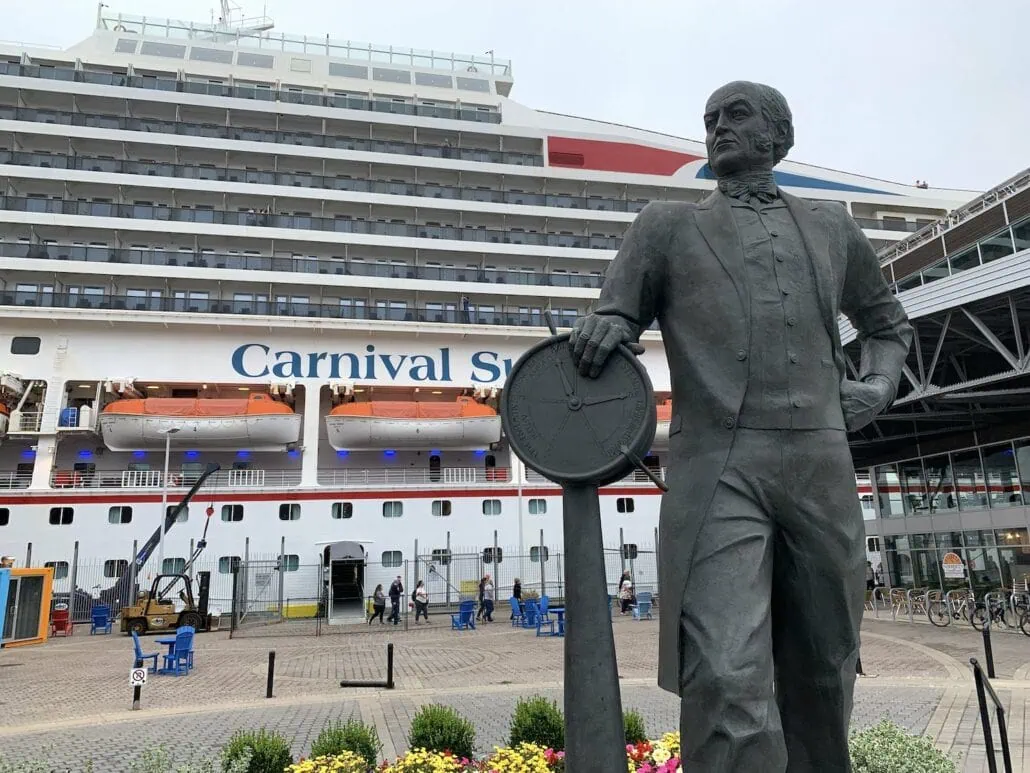 A statue of a man holding a steering wheel commands the foreground at Halifax Cruise Port, while the majestic cruise ship "Carnival Sunshine" is docked behind. People are leisurely walking and sitting, exploring what to do in this vibrant port area.