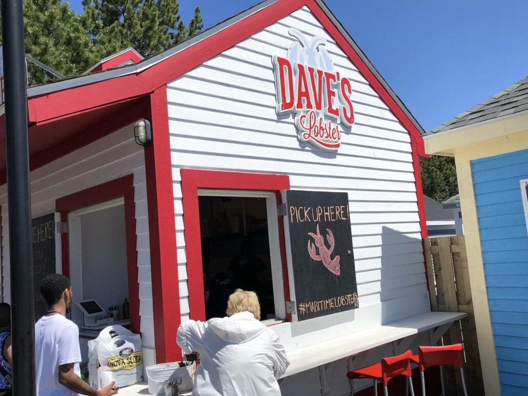 A small white and red building near Halifax Cruise Port proudly displays a sign reading "Dave's Lobster." At the front counter with stools, visitors enjoy docked activities as they place their orders. A "Pick Up Here" sign, adorned with a lobster illustration, stands out while trees sway in the background.