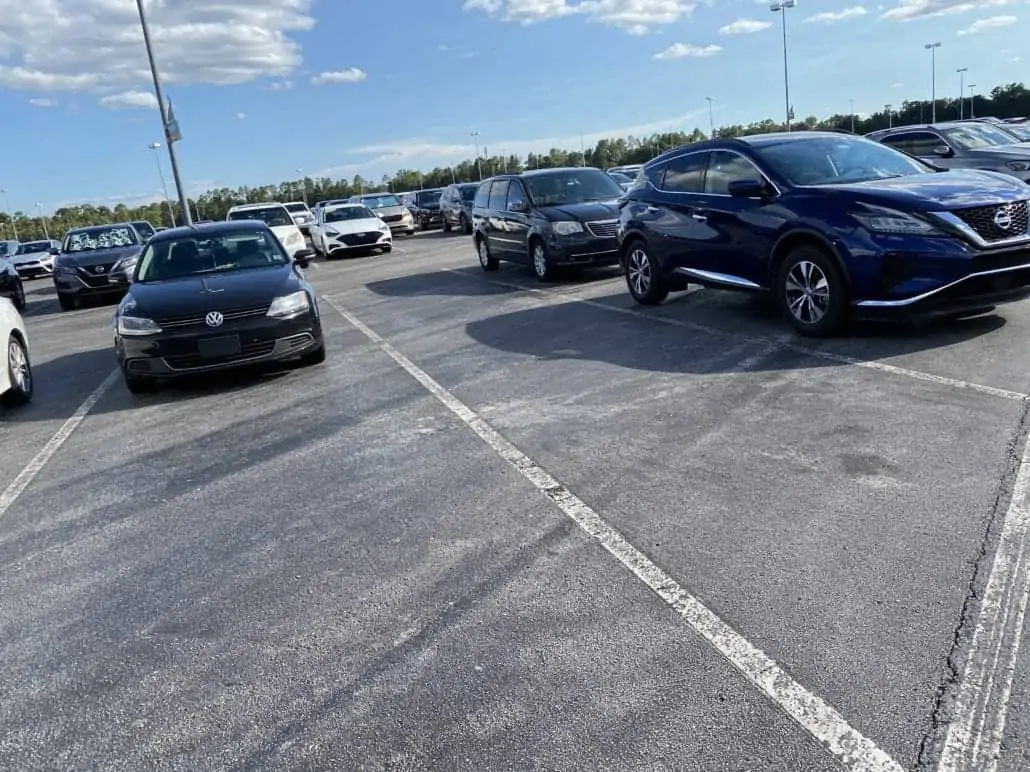 A parking lot filled with multiple cars under a clear sky. The cars include a dark-colored Volkswagen on the left and a dark-colored Nissan on the right. Visitors, perhaps fresh from Disney's Animal Kingdom, fill the mostly full lot, with other vehicles visible in the background.
